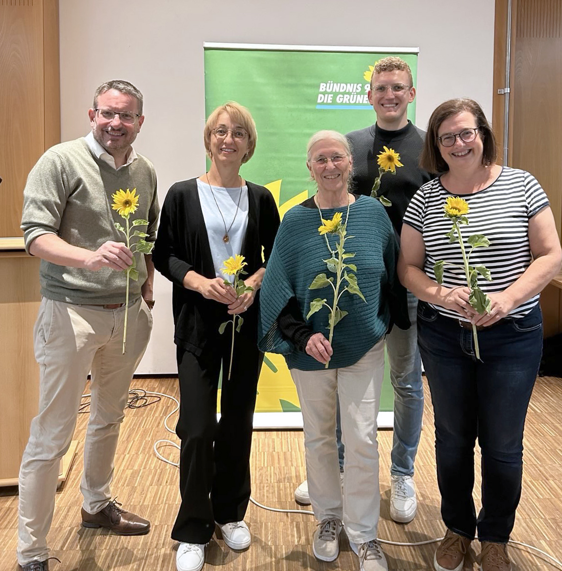 Der neue Vorstand des Kreisverbands Bündnis 90/Die Grünen in Roth steht lächelnd zusammen und hält jeweils eine Sonnenblume in den Händen. Zu sehen sind: Martin Mändl, Tanja Josche, Henrik Schmidt-Heck, Ursula Burkhardt und Eva Neubert. Im Hintergrund ist ein grünes Banner der Partei zu sehen.