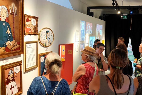Das Bild zeigt eine Gruppe von Menschen, die eine Ausstellung vom Burgmuseum Abenberg besuchen. Die Ausstellung besteht aus verschiedenen illustrierten Bildern, die historische Figuren darstellen. Ein großes Porträt zeigt einen Mann mit grauem Haar und einem blauen Gewand, der neben zwei Kerzen sitzt und ein Pergament hält. Darunter steht in einer Sprechblase: "Ich war im Rat des Königs". Die Besucher betrachten die Ausstellung interessiert, einige lesen die Beschreibungen zu den Bildern. Die Wände sind mit weiteren Illustrationen und Texten dekoriert. Es gibt auch eine interaktive Ecke mit einem orangefarbenen Rahmen. Die Atmosphäre wirkt lebhaft und bildungsorientiert.