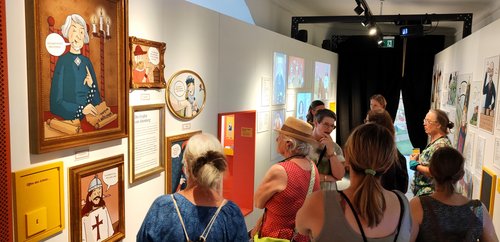 Das Bild zeigt eine Gruppe von Menschen, die eine Ausstellung vom Burgmuseum Abenberg besuchen. Die Ausstellung besteht aus verschiedenen illustrierten Bildern, die historische Figuren darstellen. Ein großes Porträt zeigt einen Mann mit grauem Haar und einem blauen Gewand, der neben zwei Kerzen sitzt und ein Pergament hält. Darunter steht in einer Sprechblase: "Ich war im Rat des Königs". Die Besucher betrachten die Ausstellung interessiert, einige lesen die Beschreibungen zu den Bildern. Die Wände sind mit weiteren Illustrationen und Texten dekoriert. Es gibt auch eine interaktive Ecke mit einem orangefarbenen Rahmen. Die Atmosphäre wirkt lebhaft und bildungsorientiert.