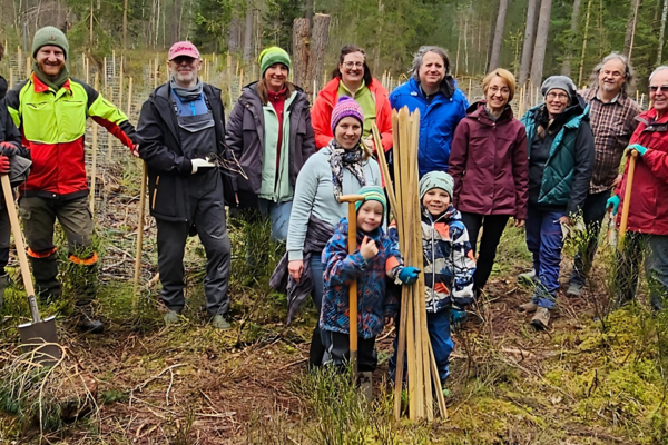 Ein Gruppe von Freiwilligen, unterschiedlichen Alters und unterschiedlicher Geschlechts, posiert lächelnd in einem Waldstück für ein Foto. Sie sind mit Pflanzwerkzeugen und Schutzausrüstung ausgestattet, bereit für eine Baumpflanzaktion. Einige Teilnehmer halten Schaufeln und junge Bäume, die zum Einpflanzen bereit sind. Es scheint ein kühler Tag zu sein, da alle warme Outdoor-Kleidung tragen, und die Stimmung wirkt fröhlich und engagiert.