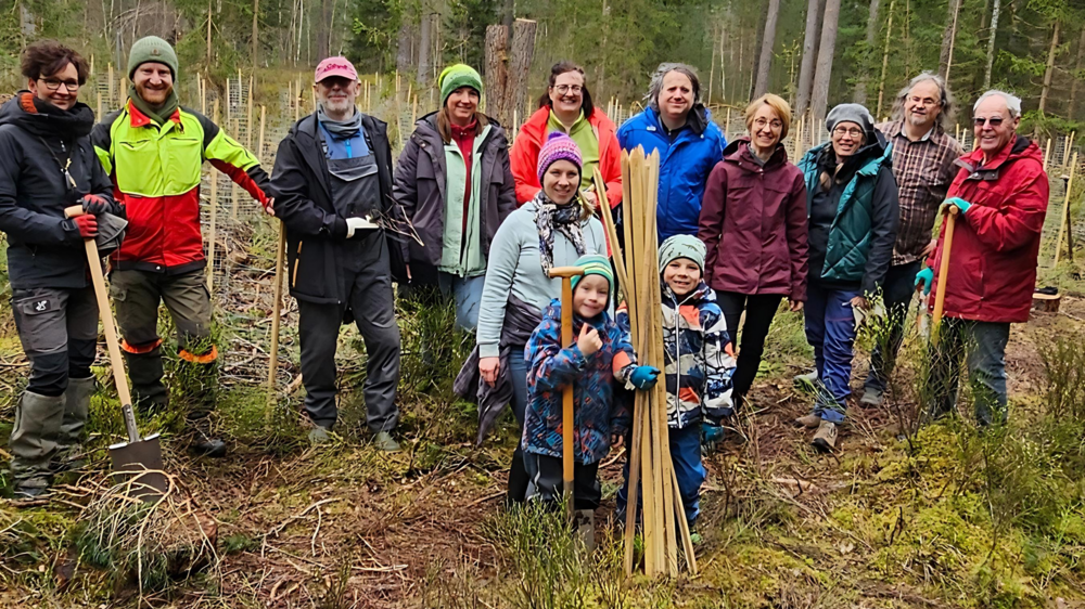 Ein Gruppe von Freiwilligen, unterschiedlichen Alters und unterschiedlicher Geschlechts, posiert lächelnd in einem Waldstück für ein Foto. Sie sind mit Pflanzwerkzeugen und Schutzausrüstung ausgestattet, bereit für eine Baumpflanzaktion. Einige Teilnehmer halten Schaufeln und junge Bäume, die zum Einpflanzen bereit sind. Es scheint ein kühler Tag zu sein, da alle warme Outdoor-Kleidung tragen, und die Stimmung wirkt fröhlich und engagiert.