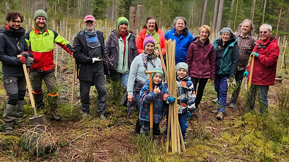 Ein Gruppe von Freiwilligen, unterschiedlichen Alters und unterschiedlicher Geschlechts, posiert lächelnd in einem Waldstück für ein Foto. Sie sind mit Pflanzwerkzeugen und Schutzausrüstung ausgestattet, bereit für eine Baumpflanzaktion. Einige Teilnehmer halten Schaufeln und junge Bäume, die zum Einpflanzen bereit sind. Es scheint ein kühler Tag zu sein, da alle warme Outdoor-Kleidung tragen, und die Stimmung wirkt fröhlich und engagiert.
