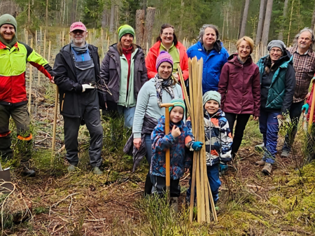 Ein Gruppe von Freiwilligen, unterschiedlichen Alters und unterschiedlicher Geschlechts, posiert lächelnd in einem Waldstück für ein Foto. Sie sind mit Pflanzwerkzeugen und Schutzausrüstung ausgestattet, bereit für eine Baumpflanzaktion. Einige Teilnehmer halten Schaufeln und junge Bäume, die zum Einpflanzen bereit sind. Es scheint ein kühler Tag zu sein, da alle warme Outdoor-Kleidung tragen, und die Stimmung wirkt fröhlich und engagiert.